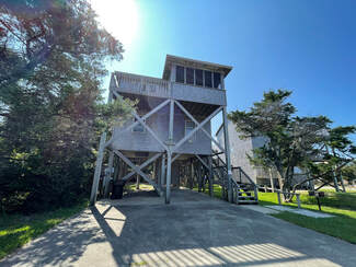 Two-story wooden house on stilts with a driveway underneath, surrounded by trees, under a clear blue sky.