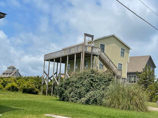 Light green two-story house with a large elevated wooden deck and exterior stairs, surrounded by greenery.