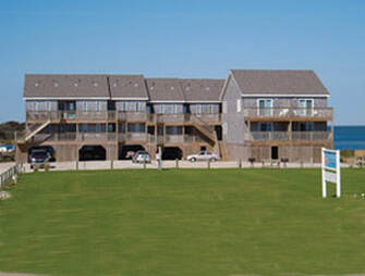 A row of wooden beachfront townhouses with balconies, parked cars underneath, and a grassy lawn in front.