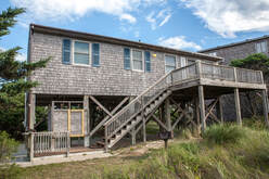 Elevated beach house with wood siding, exterior staircase, and porch, set in a grassy area under a partly cloudy sky.