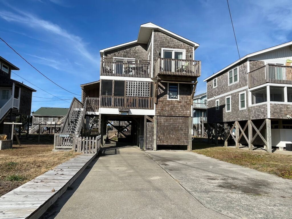 Two-story beach house on stilts with wooden exterior, multiple balconies, and a paved driveway under a blue sky.