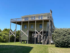 Two-story house on tall stilts with a large upper deck, set on a grassy lawn under a clear blue sky.