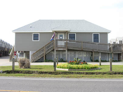 Single-story gray house with a ramp, raised foundation, and small flower bed in front, viewed from across the street.