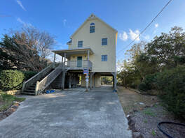 A three-story yellow house on stilts with a large driveway and outdoor staircase, surrounded by trees and bushes.