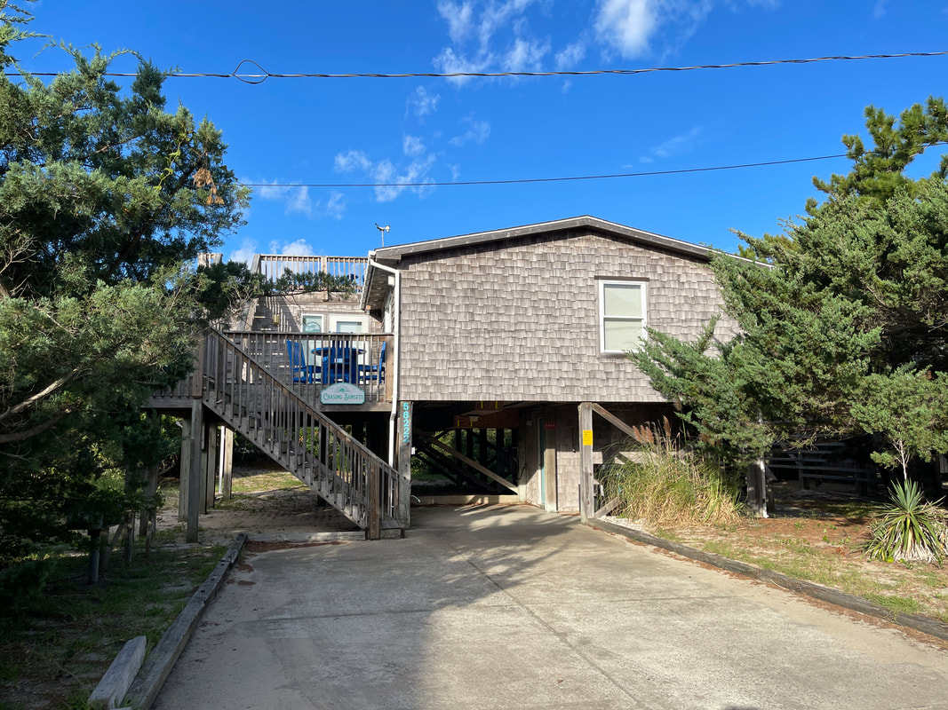 A wooden beach house on stilts with a driveway, surrounded by trees, under a blue sky with scattered clouds.
