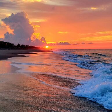 Bright orange sunset over a sandy beach with waves rolling onto the shore under a partly cloudy sky.