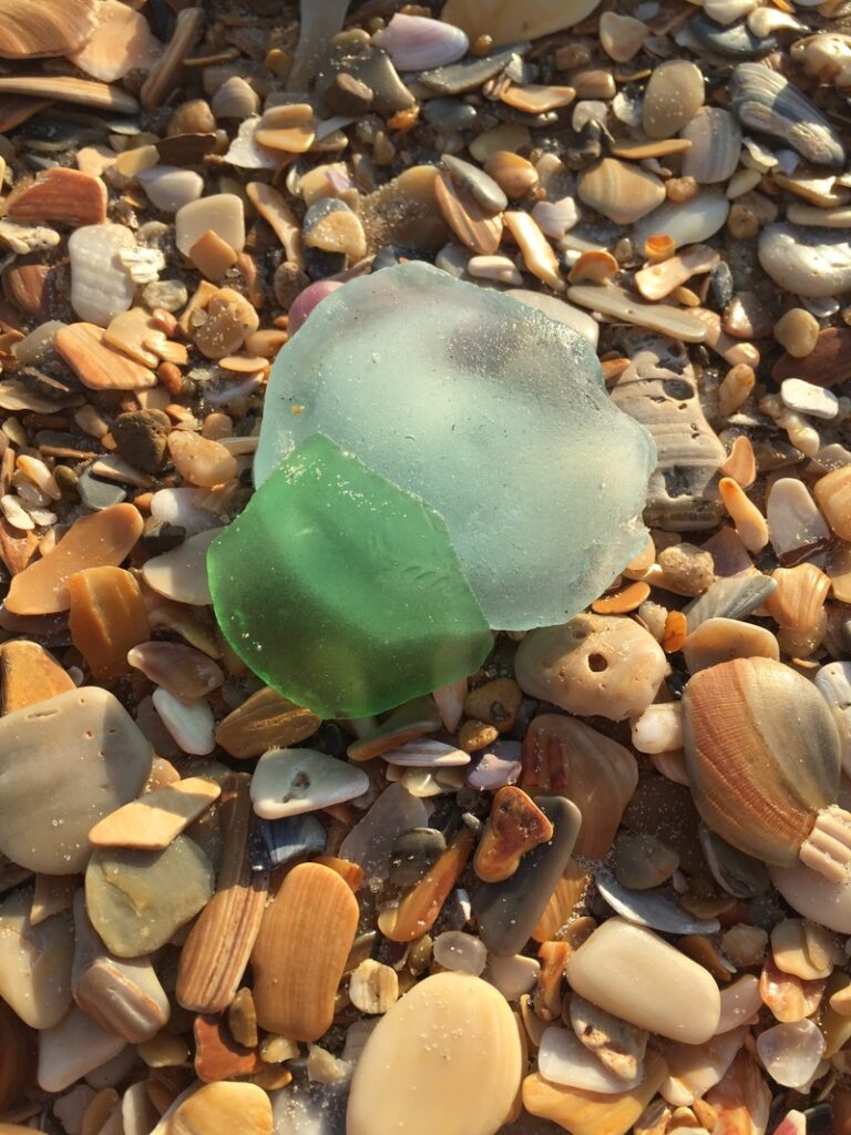 Two pieces of sea glass, one green and one light blue, rest among assorted seashells on a sandy beach.