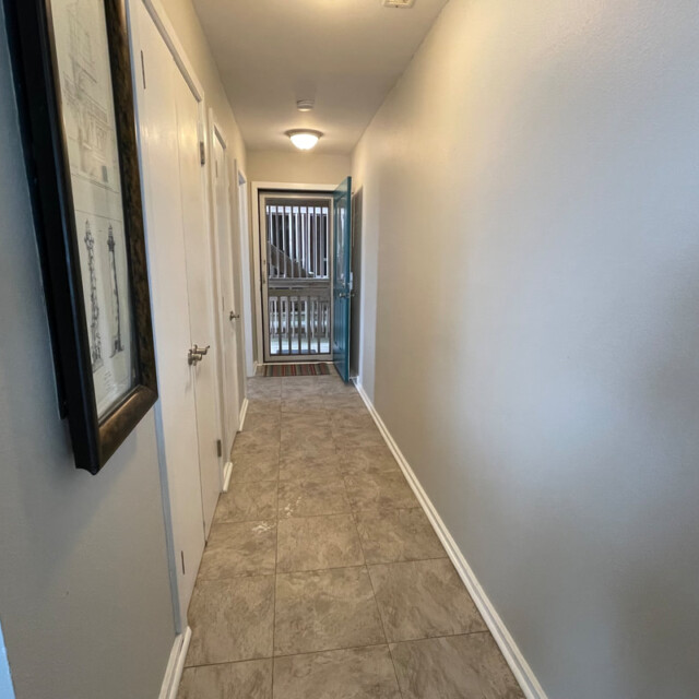 A narrow hallway with tile floor, white walls, doors on the left, and an open door leading outside at the end.