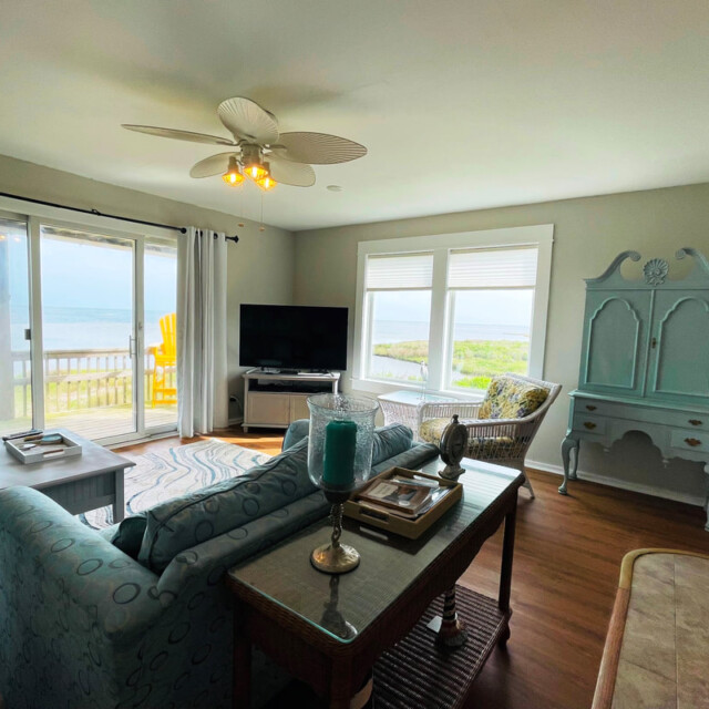 Bright living room with large windows, a TV, blue furniture, and a view of a balcony overlooking water and greenery.