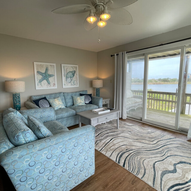 Living room with blue patterned sofas, sea-themed decor, and large windows overlooking a lake with a balcony.