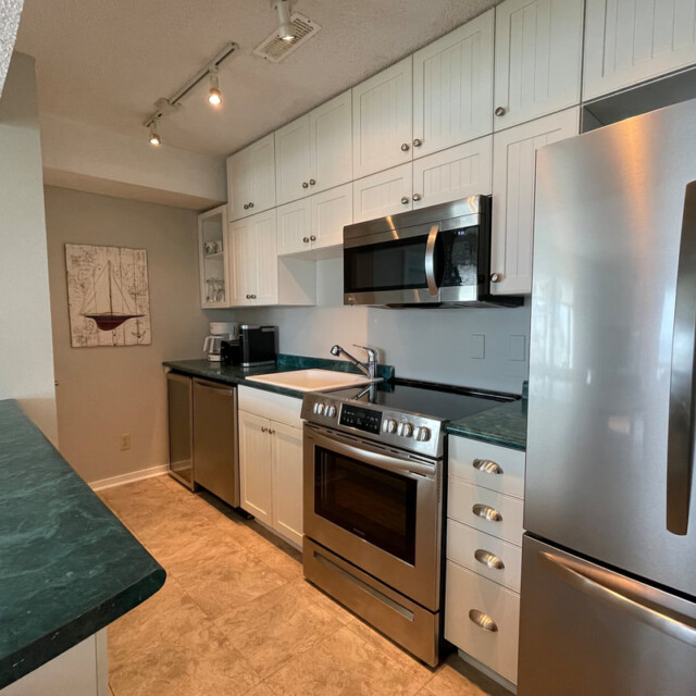 Modern kitchen with stainless steel appliances, white cabinets, green countertops, and beige tile flooring.