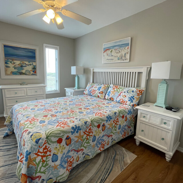 Bedroom with a king bed, coastal-themed bedding, two nightstands, two lamps, and seaside wall art.