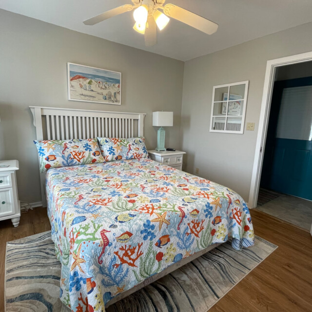 Bedroom with a queen bed, colorful ocean-themed bedding, two nightstands, and a ceiling fan.