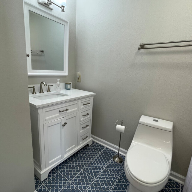 Small bathroom with a white vanity, mirror, modern toilet, towel rack, and blue patterned tile floor.
