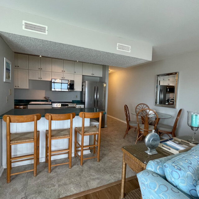 Open kitchen with bar stools, adjacent dining area, and part of a living room with a blue-patterned couch.