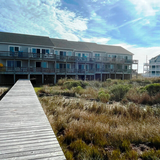 A wooden boardwalk leads to a two-story beach house with balconies, surrounded by grass and clear blue sky.