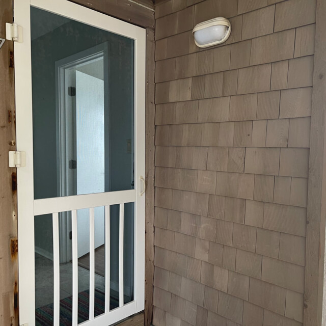 Brown shingled exterior with a white screen door, outdoor light, and a view into an interior hallway.