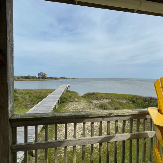 Wooden porch with yellow chair overlooks a boardwalk leading to a calm bay under a partly cloudy sky.
