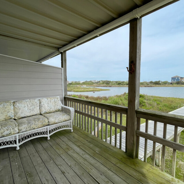 Covered porch with a wicker sofa overlooks a wooden walkway leading to water and grassy landscape under a clear sky.