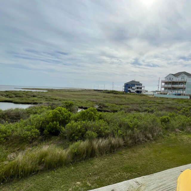 View of grassy wetlands with houses in the distance under a cloudy sky, seen from a porch with a yellow chair.