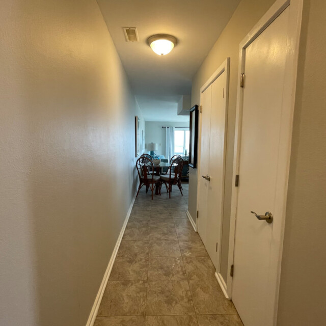 A narrow hallway with tiled floor leads to a dining area with a table and chairs, lit by a ceiling light.