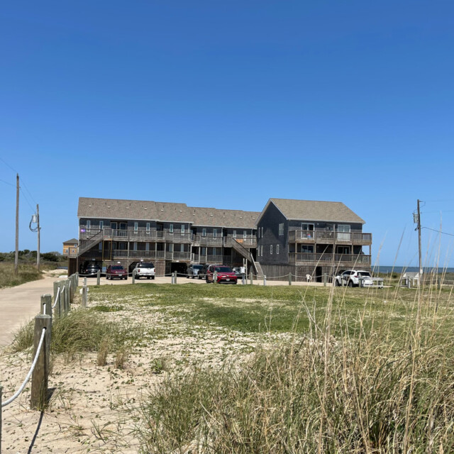 Two-story beachfront building with parked cars, grassy sand dunes, a paved walkway, and clear blue sky.