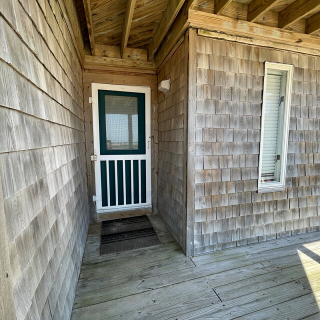 Wooden porch with shingle siding, a white-trimmed screen door, two windows, and a doormat in front of the entrance.