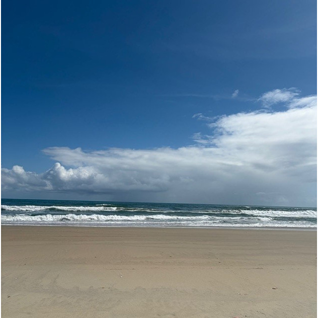 Sandy beach with gentle waves under a clear blue sky and scattered clouds.