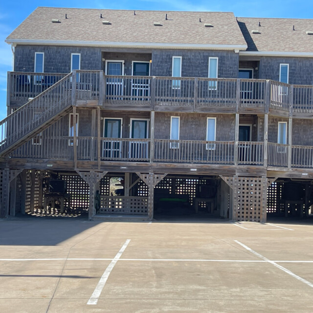 Three-story wooden beachfront building with open parking spaces below and balconies on each level, under a clear sky.