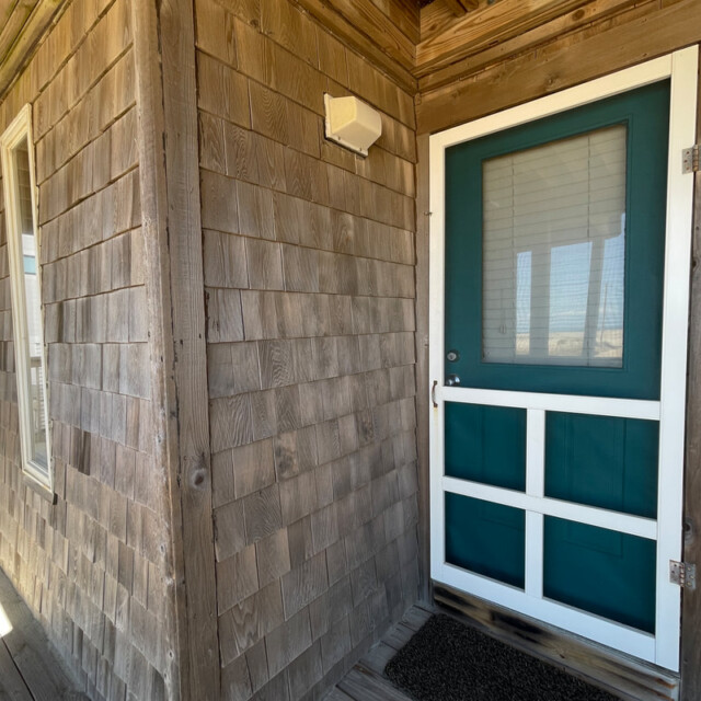 Wooden exterior with a teal and white door labeled “1,” window, covered porch, and doormat in front.