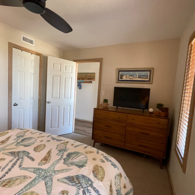 Bedroom with a starfish-patterned bedspread, dresser with TV, open door, and window with blinds.