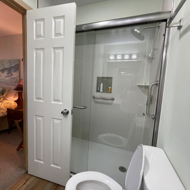 Open bathroom door reveals a glass shower, white toilet, and glimpse of a bedroom with floral bedding in background.