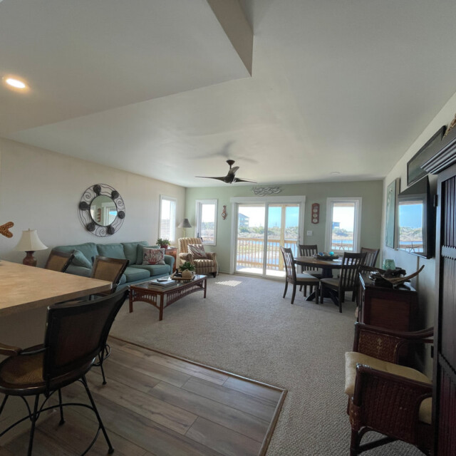 Bright living and dining area with coastal decor, large windows, and balcony overlooking the ocean.