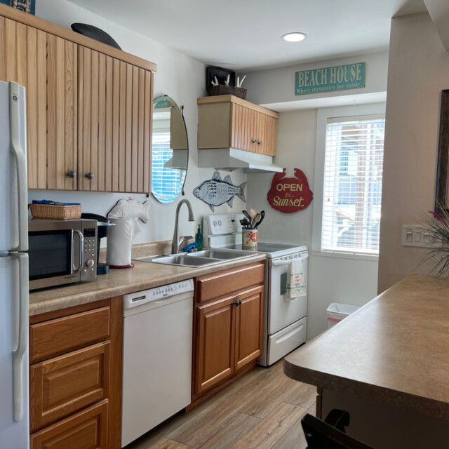 Small kitchen with white appliances, wood cabinets, beach decor, and sunlight coming through a window above the sink.