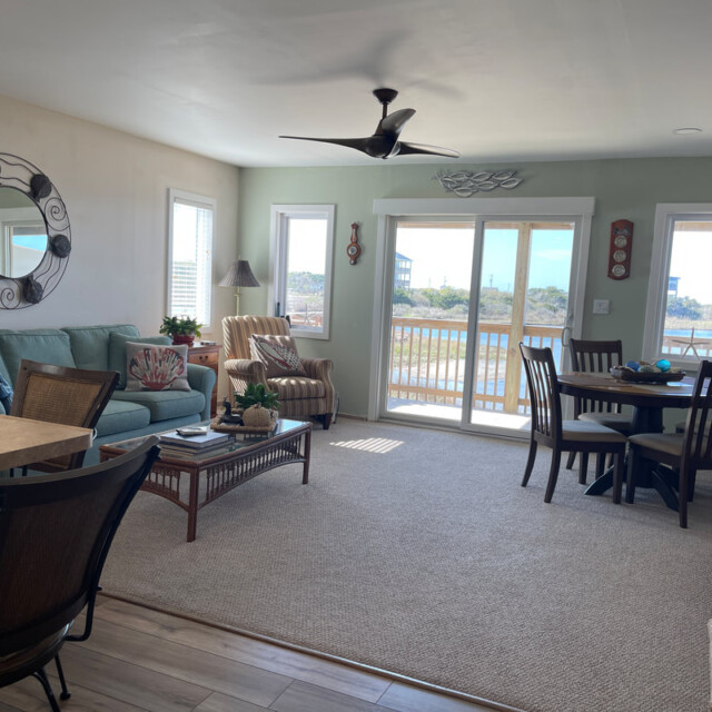 Bright living and dining area with large windows, light furniture, and a balcony overlooking water and trees.