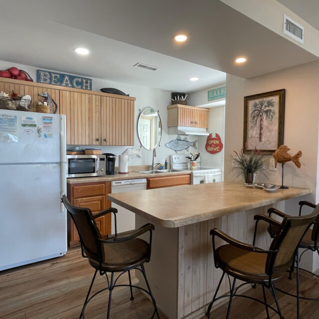 Small beach-themed kitchen with wooden cabinets, white appliances, bar counter, and three chairs.