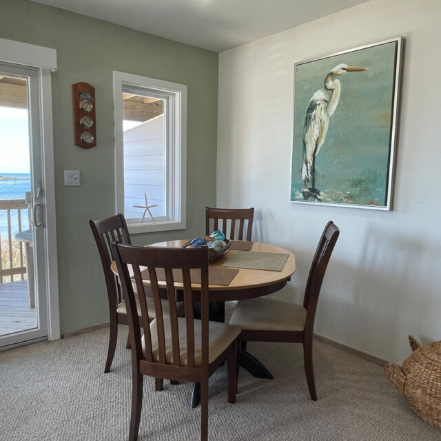 Small dining area with four chairs, a round table, heron artwork, and a view of a porch and water through glass doors.