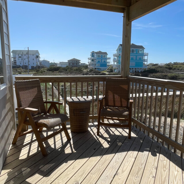 Wooden deck with two wicker chairs and a table, overlooking houses and dunes under a clear sky.