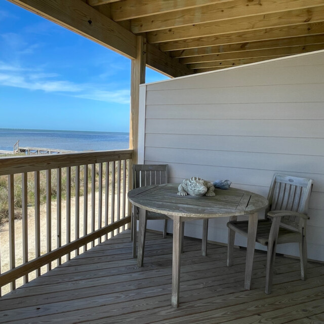 Wooden deck with two chairs and a round table overlooks the beach and ocean under a clear blue sky.