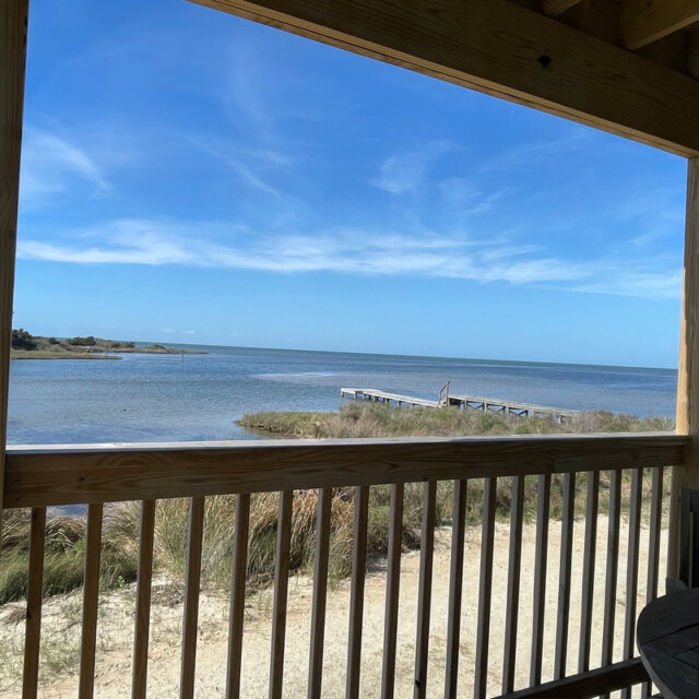 View of a calm bay and dock seen from a covered porch with wooden railings and outdoor furniture under a clear sky.