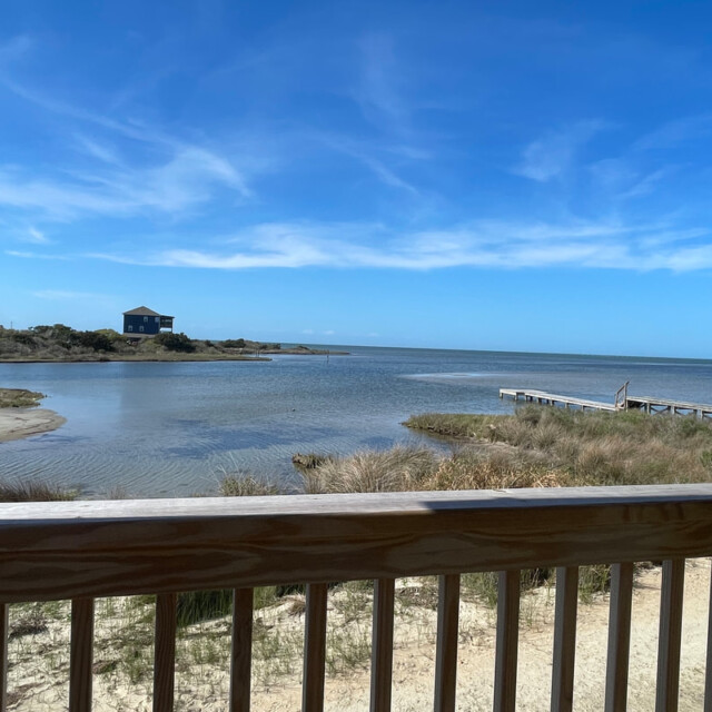 View of a calm bay with a dock, grassy shore, and a blue house in the distance under a clear sky.