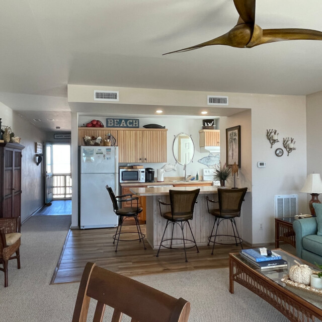 Open kitchen and living area with coastal decor, wicker furniture, and a large ceiling fan in a beach-themed home.