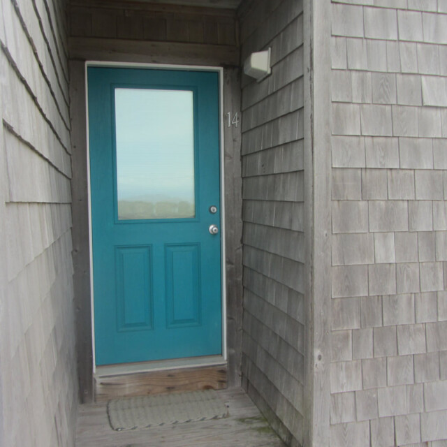 A teal door with a window, labeled 14, set in a shingle-style house with a doormat in front.