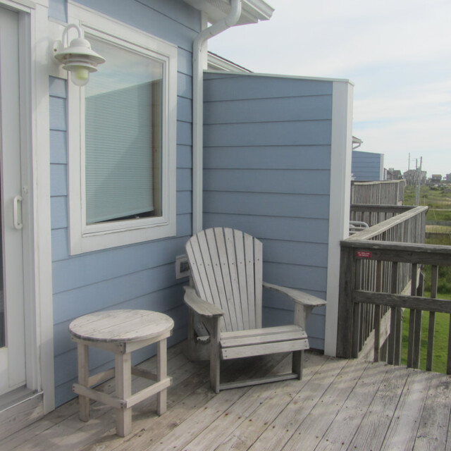 Wooden chair and round table on a wooden deck outside a blue house, with grassy area and houses in the background.
