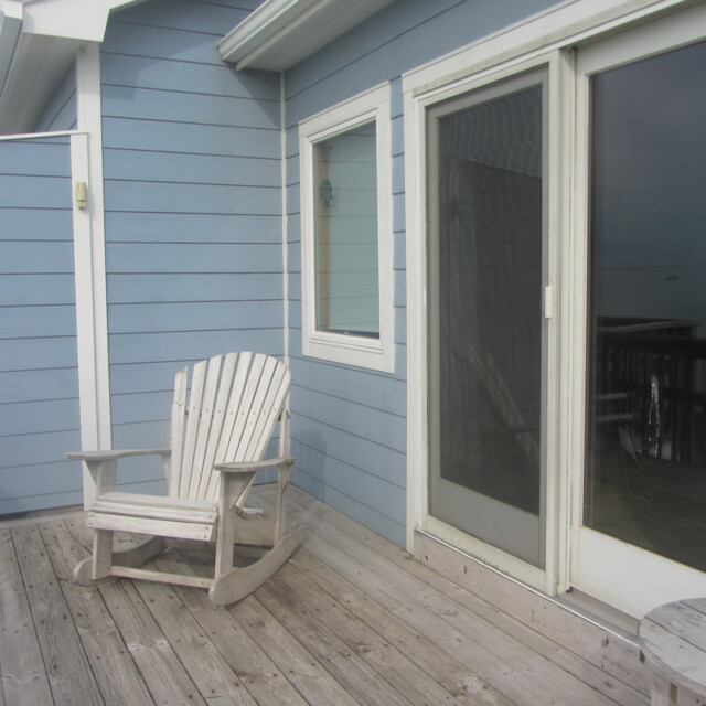 Wooden rocking chair sits on a wooden deck outside a blue house with sliding glass doors and a white-framed window.