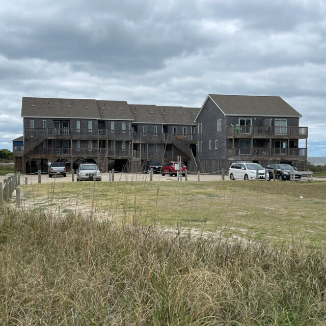 Three gray-shingled beach houses with parked cars on a cloudy day, viewed from a grassy area in the foreground.