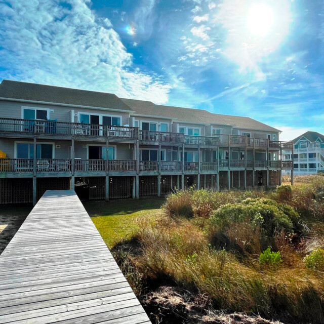 Two-story beachfront building with wooden walkway, grassy dunes, and clear blue sky with sun overhead.