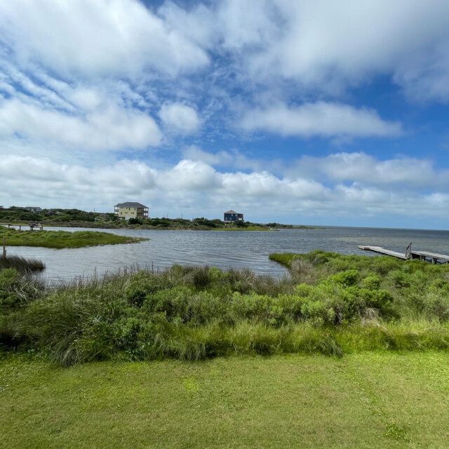 Grassy shoreline with docks, calm water, and houses in the distance under a partly cloudy blue sky.