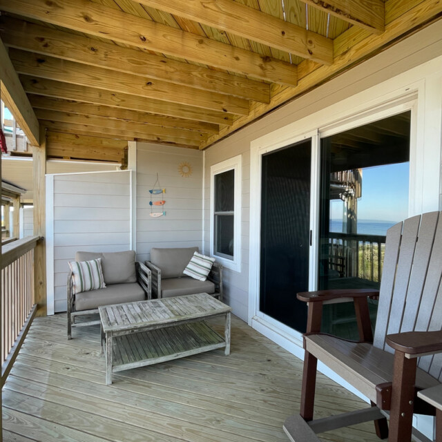 Covered wooden balcony with seating, a small table, decorative wall art, and sliding glass doors to the interior.