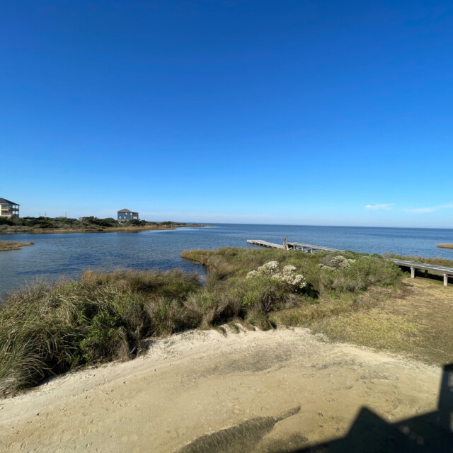Sandy shore with grassy dunes, a wooden walkway, and two houses near a calm body of water under a clear blue sky.